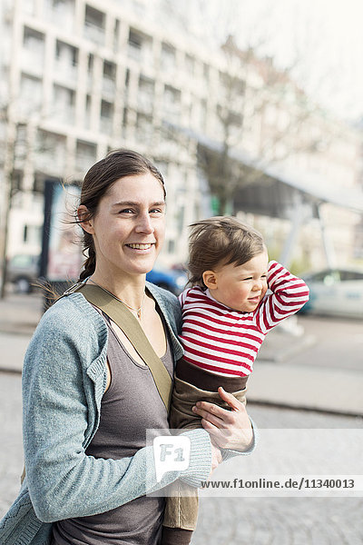 Porträt einer glücklichen Mutter die ein kleines Mädchen in der Stadt trägt.
