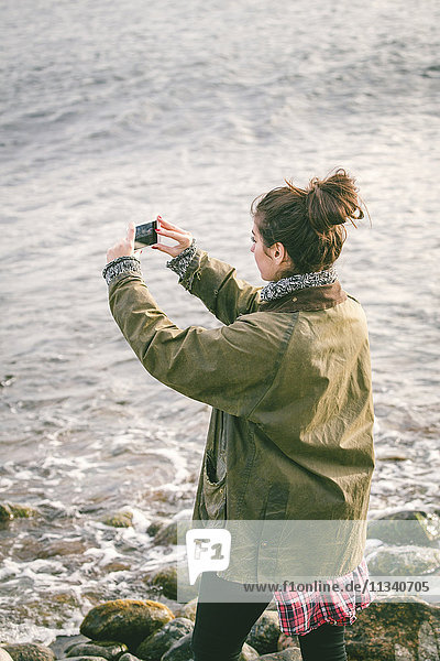 Frau fotografiert das Meer  während sie am Strand steht.