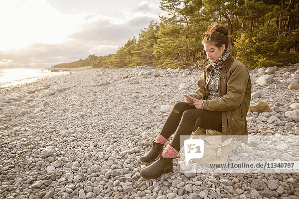Woman sitting on rock with bag while using mobile phone at beach