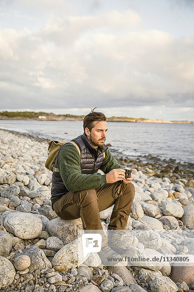 Mann auf Felsen sitzend mit Kamera am Strand gegen den Himmel