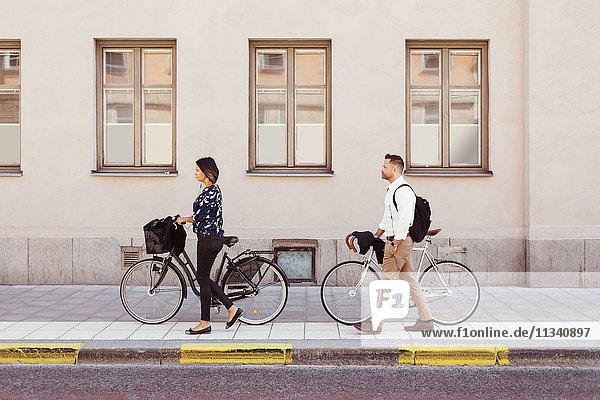 Seitenansicht von Geschäftskollegen  die mit Fahrrädern auf dem Bürgersteig laufen