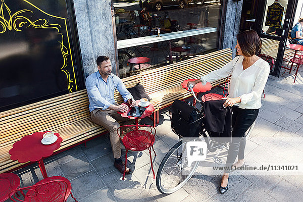 Businesswoman with bicycle talking to male colleague sitting at sidewalk cafe