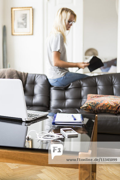 Technik und Buch am Tisch mit Teenager-Lesedokument beim Sitzen auf dem Sofa im Wohnzimmer