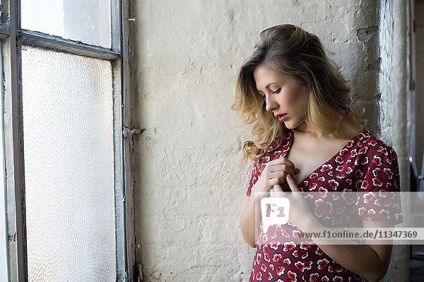 Beautiful young woman in red dress by the window hands over heart.