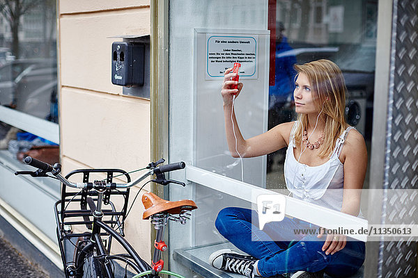 Junge Frau mit Handy und Ohrhörern hinter einer Fensterscheibe