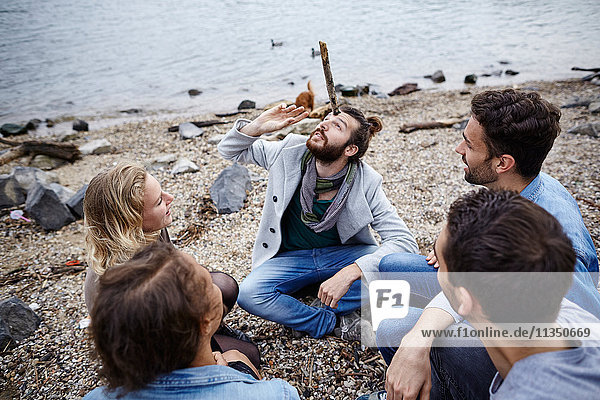 Man sitting with friends at the riverside balancing a stick on his head