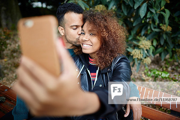 Lächelndes junges Paar auf einer Parkbank macht ein Selfie