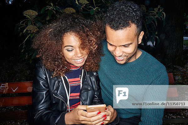 Smiling young couple on park bench looking at cell phone