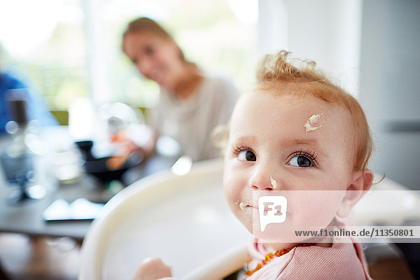 Baby mit Essensresten im Gesicht und Mutter im Hintergrund