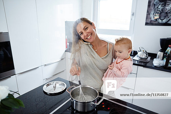 Smiling mother with baby girl cooking and telephoning