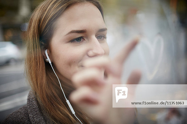 Junge Frau mit Ohrhörern zeichnet ein Herz in eine Glasscheibe