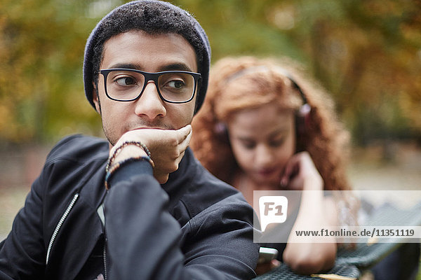 Ernster junger Mann auf einer Parkbank mit Freundin im Hintergrund