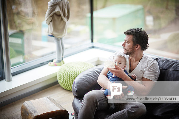 Vater gibt seinem Baby das Fläschchen auf der Couch mit Tochter am Fenster