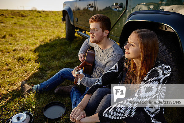Junges Paar sitzt auf einer Wiese neben einem Auto und macht Musik