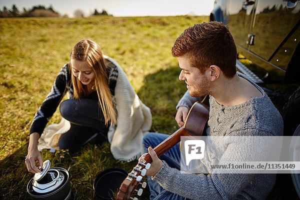 Junges Paar sitzt auf einer Wiese neben einem Auto und einem Campingkocher und macht Musik