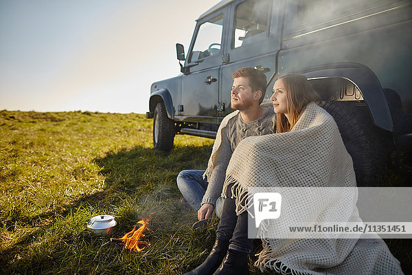 Junges Paar sitzt auf einer Wiese neben einem Auto und Campingkocher