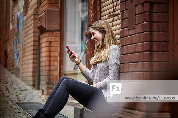 Lachende junge Frau sitzt auf der Türschwelle und schaut auf ihr Handy