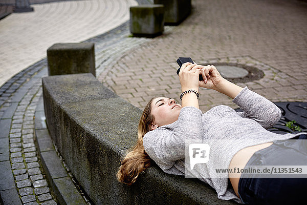 Junge Frau liegt auf einer Mauer und schaut auf ihr Handy