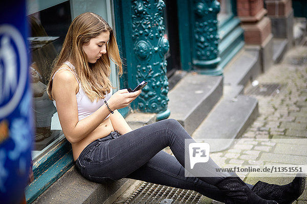 Junge Frau sitzt auf der Türschwelle und schaut auf ihr Handy