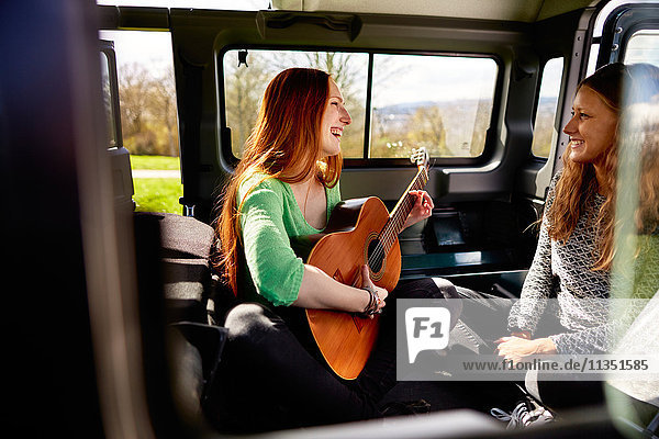 Smiling young woman looking at female friend playing guitar in car