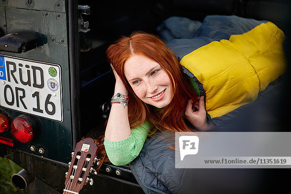 Smiling young woman in sleeping bag lying in off-road vehicle