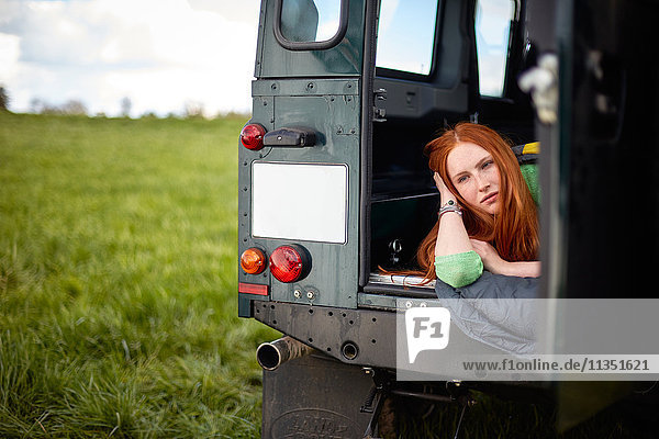 Junge Frau mit Schlafsack liegt in einem Geländewagen auf einer Wiese