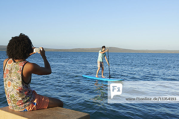 Junger Mann fotografiert Freund beim Paddeln auf dem sonnigen Sommermeer
