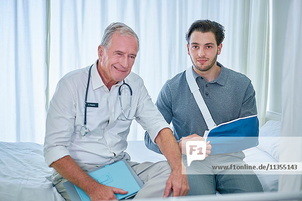 Arzt und Mann mit Arm im Hebegurt auf Krankenhausbett sitzend