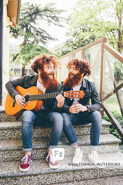 Junge männliche Hipster-Zwillinge mit roten Bärten sitzen auf einer Treppe und spielen Gitarre
