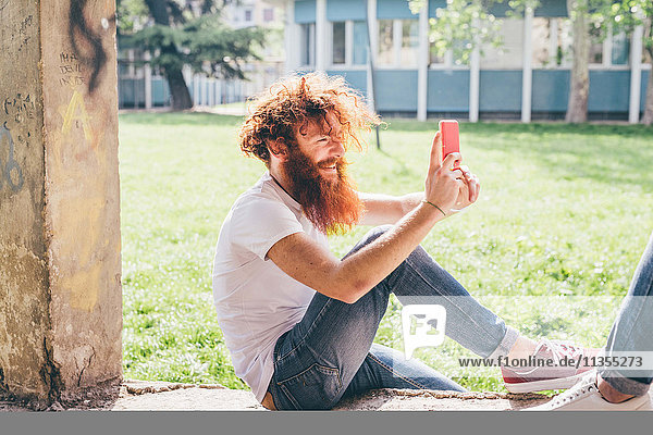 Junger männlicher Hipster mit roten Haaren und Bart fotografiert Freund auf Smartphone im Park