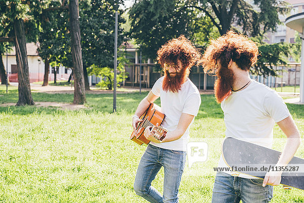 Young male hipster twins with red hair and beards strolling in park playing guitar