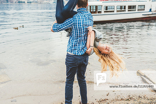Junger Mann trägt seine Freundin kopfüber am Seeufer,  Comer See,  Italien