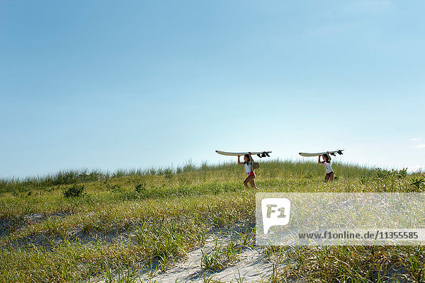 Zwei junge Mädchen gehen mit Surfbrettern auf Sanddünen