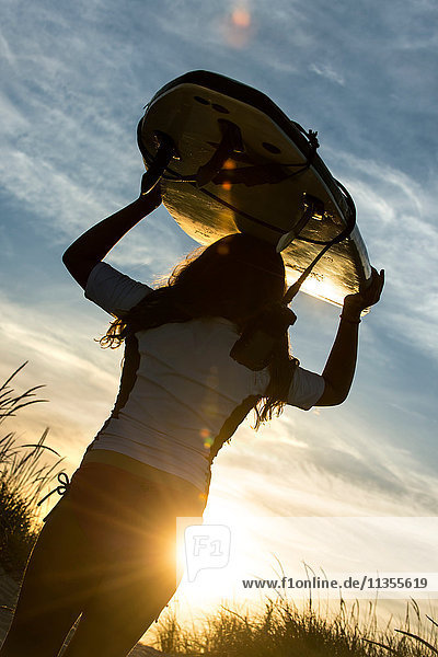 Junges Mädchen am Strand  ein Surfbrett tragend  bei Sonnenuntergang  Rückansicht