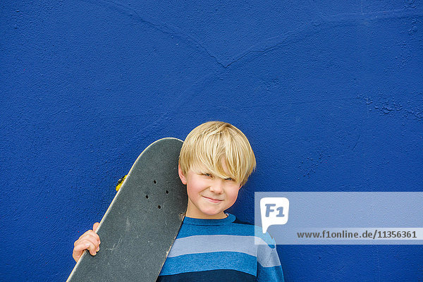 Porträt eines süßen Jungen  der ein Skateboard auf der Schulter trägt  vor einer blauen Wand