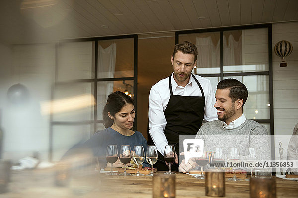 Man wearing apron standing by business people while pointing at wineglasses on table