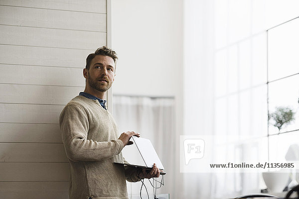 Businessman holding laptop while standing against wall in office