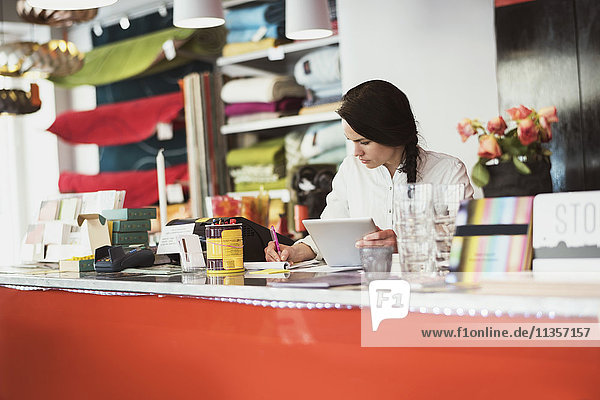 Besitzerin mit digitalem Tablett beim Schreiben auf Notizblock an der Kasse im Geschäft