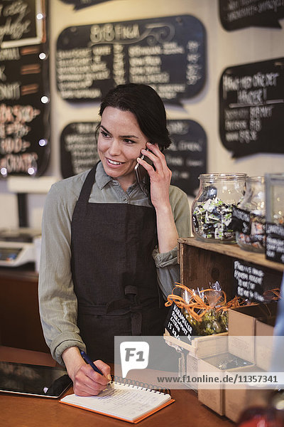 Lächelnde Besitzerin mit Telefon beim Schreiben im Notizblock am Ladentisch