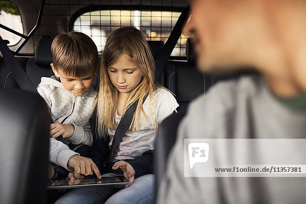 Sibling using digital tablet while sitting in car with father in foreground