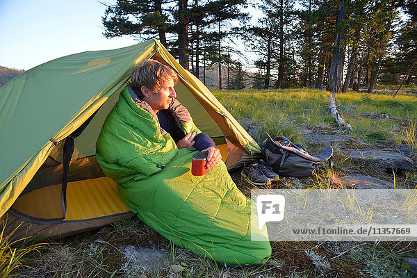 Männlicher Camper  der im Schlafsack sitzend den Sonnenuntergang am Midnight Ridge beobachtet  Colville National Forest  Bundesstaat Washington  USA