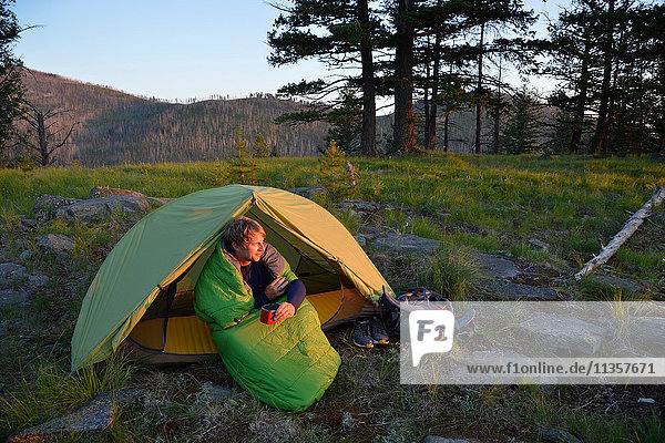 Männlicher Camper  der im Schlafsack sitzend den Sonnenuntergang am Midnight Ridge beobachtet  Colville National Forest  Bundesstaat Washington  USA
