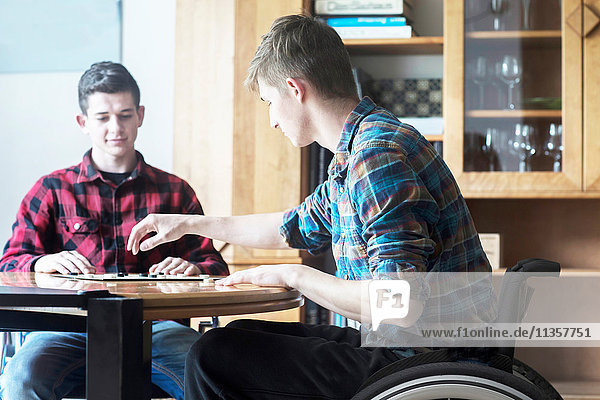 Junger Mann im Rollstuhl beim Damespiel mit einem Freund in der Küche