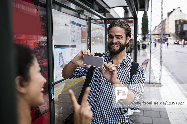 Lächelnder Mann fotografiert Freundin durch Smartphone an der Bushaltestelle in der Stadt