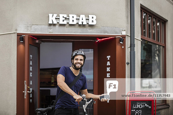 Portrait of smiling man with bicycle holding smart phone while standing against restaurant in city
