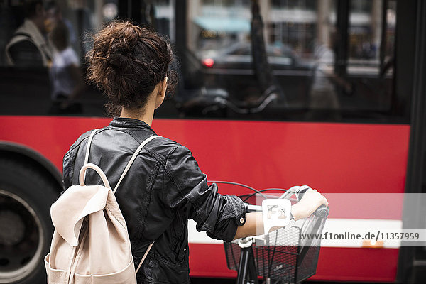 Rear view of woman with bicycle carrying backpack while standing against red bus in city