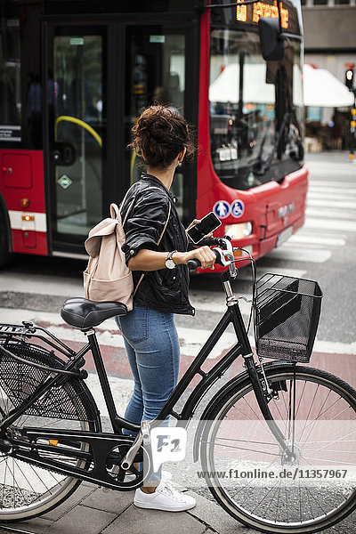 Volle Länge der Frau mit Fahrrad  die den Bus betrachtet  während sie auf der Stadtstraße steht.