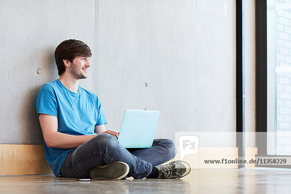 Junger männlicher Student mit Laptop auf dem Boden sitzend an einer Hochschule