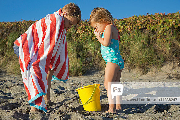 Boy wrapped in towel looking down at toy bucket with sister on beach  Blowing Rocks Preserve  Jupiter Island  Florida  USA