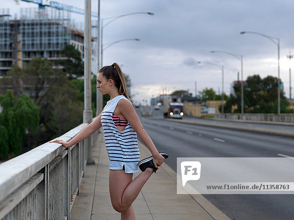 Young female runner stretching legs on highway bridge at dawn